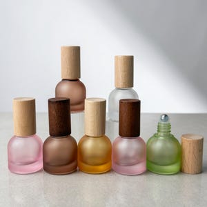May include: A collection of seven small, round, frosted glass bottles with wooden caps. The bottles come in various colors including pink, brown, amber, and green. One green bottle is shown without its cap, revealing a metal roller ball top.