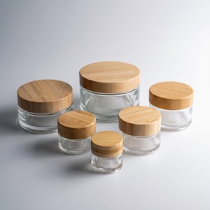 May include: A selection of clear glass jars with bamboo lids. The jars are of varying sizes, with different diameters. The lids are made of light-coloured bamboo, providing a natural contrast with the transparent glass. The jars are displayed on a white surface.