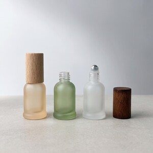 May include: Two frosted glass roller bottles with wooden caps. One bottle is light peach with a light wood cap, and the other is clear frosted with a metal roller ball and a dark wood cap. The bottles have a rounded base and threaded necks.