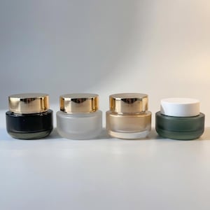 Op de afbeelding: Vier kleine cosmetische potjes met goudkleurige deksels. De potjes zijn in verschillende kleuren: zwart, mat, lichtgoud en groen. De potjes staan op een wit oppervlak.