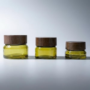 Könnte beinhalten: Drei Glasbehälter mit olivgrünen Körpern und dunkelbraunen Holzdeckeln. Die Gläser sind unterschiedlich groß und bilden eine abgestufte Anzeige. Die Gläser dienen wahrscheinlich der Aufbewahrung oder Dekoration.