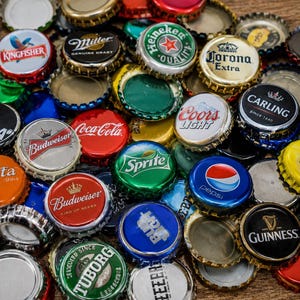 Może przedstawiać: Kolekcja różnorodnych metalowych kapsli z logotypami różnych marek, w tym Bud Light, Kingfisher, Miller, Heineken, Corona Extra, Soda, Coca-Cola, Coors Light, Carling, Budweiser, Fanta, Sprite, Pepsi, Schweppes, Tuborg oraz Guinness.