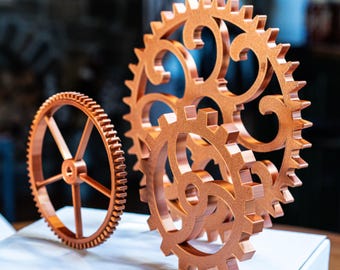 以下が含まれることがあります： 銅色の歯車モデルが2つ。1つはスポークと歯付きリムが付いたソリッドホイールです。もう1つは、オープンな透かし彫りのデザインの、より大きく装飾的な歯車です。歯車は白い表面に表示されています。
