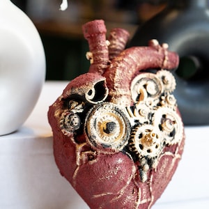 May include: A burgundy-colored anatomical heart sculpture with gold accents and intricate gear details. The steampunk-inspired art piece features a textured surface and is a unique decorative item.