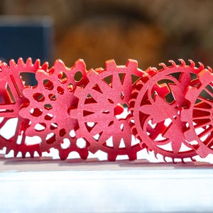 Könnte beinhalten: Reihe von sechs roten Zahnrädern mit unterschiedlichen Designs. Die Zahnräder bestehen aus einem festen Material und überlappen sich leicht. Sie werden auf einer weißen Oberfläche präsentiert. Die Zahnräder haben einen Durchmesser von ca. 10 cm.