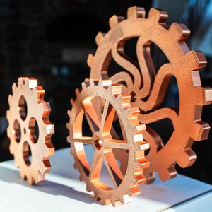 May include: Three copper-colored gear sculptures of varying sizes are displayed on a white surface. The gears have intricate designs and are arranged to showcase their mechanical aesthetic. The warm tones of the copper contrast with the neutral background.