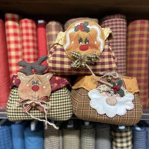 Può includere: Tre sacchetti decorativi in tessuto a tema natalizio. Le borse presentano un omino di pan di zenzero, una renna e Babbo Natale. Ogni borsa è realizzata in tessuto a quadri e ha un volto decorativo e abbellimenti. Le borse sono esposte su uno scaffale con rotoli di tessuto.