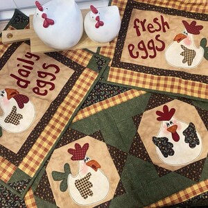 Può includere: Una collezione di tessuti da cucina a tema country con motivi di galline. Include tovagliette e un runner da tavolo con le scritte "daily eggs" e "fresh eggs". I tessuti hanno un motivo a quadri beige, verde e giallo.