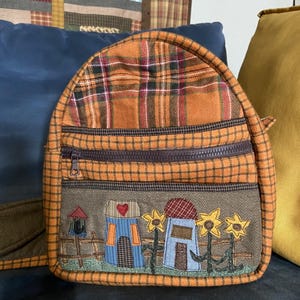Puede incluir: Una pequeña mochila a cuadros naranja con una cremallera marrón y un panel frontal decorativo. El panel presenta un diseño de patchwork de casas, girasoles y un pajarera. La mochila tiene una parte superior redondeada y una cremallera marrón.