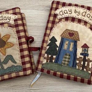 Può includere: Due diari rivestiti in tessuto con un motivo a quadri rossi e beige. Un diario presenta un'applicazione di girasole, l'altro una casa e un albero. Entrambi hanno uno striscione che dice "day by day".
