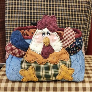 May include: A quilted fabric hen-shaped basket with a red comb, a white face, and a yellow beak. The body is made of blue denim and plaid fabric, with star-shaped feet. The basket is designed with a country theme.