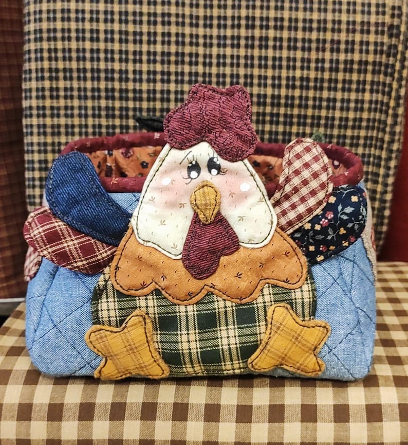 May include: A quilted fabric hen-shaped basket with a red comb, a white face, and a yellow beak. The body is made of blue denim and plaid fabric, with star-shaped feet. The basket is designed with a country theme.