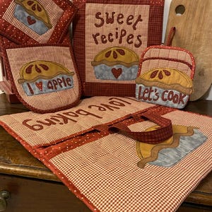 Può includere: Una collezione di tessuti da cucina a quadretti rossi e bianchi. Gli articoli includono presine, guanti da forno, un libro di ricette e una tovaglietta. Ogni articolo presenta un disegno di torta applicato con le parole "I love apple" e "Let's cook".