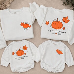 May include: Four white long-sleeved garments featuring orange pumpkin and leaf graphics. Two sweatshirts display the text DADA and OUR LITTLE PUMPKIN. Two rompers feature the text OUR LITTLE PUMPKIN and MINI, each accented with small hearts and stars.