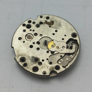 Puede incluir: Primer plano de un movimiento de reloj antiguo. El mecanismo circular plateado presenta numerosos engranajes peque&ntilde;os, tornillos y otros componentes. Se ven algunos engranajes dorados, junto con varios agujeros peque&ntilde;os. El movimiento del reloj es probablemente una pieza de repuesto o para reparaci&oacute;n.