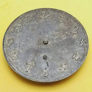 K&ouml;nnte beinhalten: Ein verwittertes, kreisf&ouml;rmiges Zifferblatt mit silber-grauer Oberfl&auml;che. Die Zahlen 1 bis 12 sind deutlich am Rand markiert. Die Zeiger zeigen auf etwa 6 Uhr. Das Zifferblatt hat ein zentrales Loch und zwei kleinere L&ouml;cher.