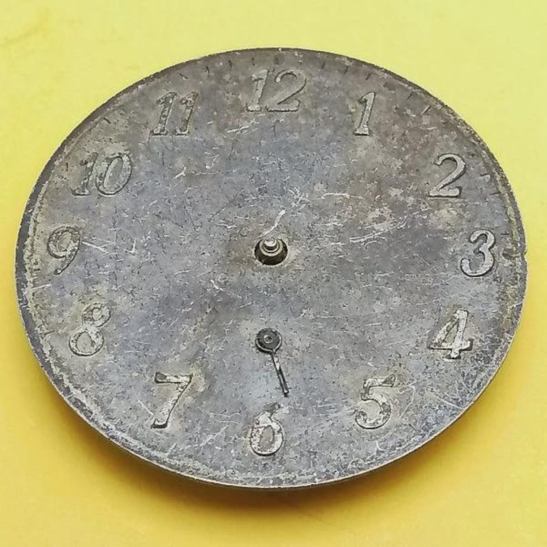 K&ouml;nnte beinhalten: Ein verwittertes, kreisf&ouml;rmiges Zifferblatt mit silber-grauer Oberfl&auml;che. Die Zahlen 1 bis 12 sind deutlich am Rand markiert. Die Zeiger zeigen auf etwa 6 Uhr. Das Zifferblatt hat ein zentrales Loch und zwei kleinere L&ouml;cher.