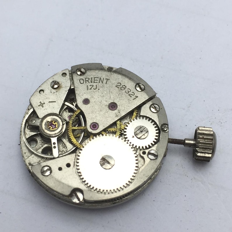 Pu&ograve; includere: Primo piano di un movimento di orologio Orient 17J argentato con ingranaggi, gioielli e una corona visibili. Il testo "ORIENT 17J" e "28321" sono stampati. Il meccanismo &egrave; una complessa disposizione di piccole parti.