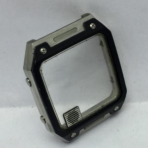 May include: A silver and black watch case with a clear, rectangular face. The case has a black bezel and is secured with small screws. A small, rectangular emblem is visible on the face.