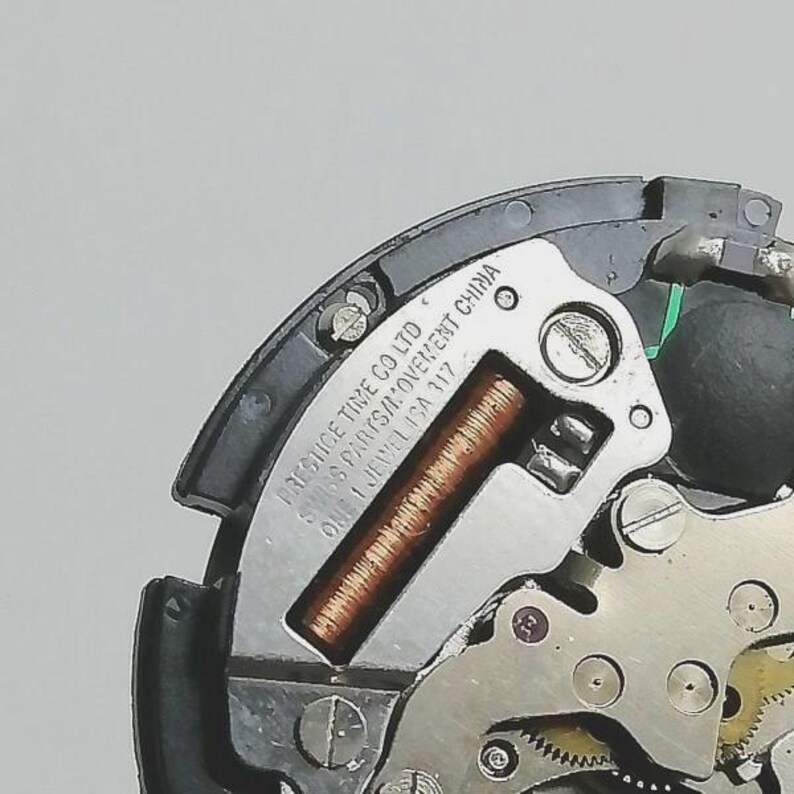 Puede incluir: Primer plano de un movimiento de reloj, con engranajes visibles, una bobina de cobre y una placa plateada. La placa est&aacute; marcada con "PRESTIGE TIME CO LTD SWISS PARTS/MOVEMENT CHINA ONE 1 JEWEL ISA 117".