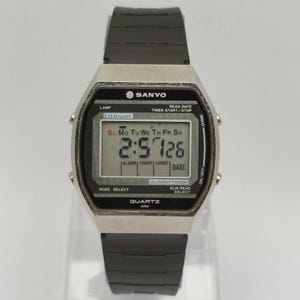 Puede incluir: Un reloj digital Sanyo vintage con una correa de goma negra. La esfera del reloj muestra la hora, la fecha y el día de la semana. El reloj tiene una caja plateada y varios botones de función. La hora mostrada es 2:57:26.