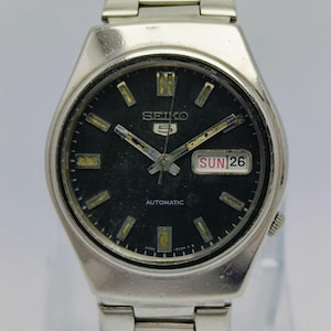May include: Silver-toned Seiko 5 automatic watch with a black dial. The watch face displays the day and date, with the day reading "SUN 26". The watch has a silver band and a classic design.