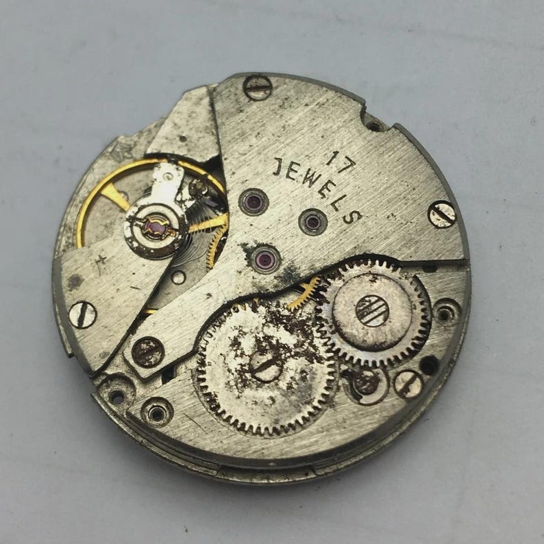 Puede incluir: Primer plano de un movimiento de reloj vintage con engranajes, tornillos y la inscripci&oacute;n "17 JEWELS". Los componentes met&aacute;licos tienen un acabado plateado con signos de desgaste. El dise&ntilde;o intrincado muestra el funcionamiento interno de un reloj mec&aacute;nico.