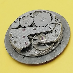 K&ouml;nnte beinhalten: Nahaufnahme eines Vintage-Uhrwerks mit komplizierten Zahnr&auml;dern und Komponenten. Die Metallteile sind silberfarben mit kleinen, roten Juwelen. Der Uhrenmechanismus ist vor einem gelben Hintergrund platziert und zeigt die detaillierte Handwerkskunst.