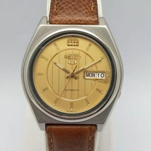 May include: A vintage Seiko 5 automatic watch with a gold-tone dial and a brown leather strap. The watch face displays the day and date, with the day reading "MON 10". The watch has a silver-tone case and a textured brown leather band.