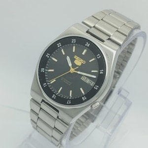 May include: A silver-toned Seiko automatic wristwatch with a black dial. The watch features gold-colored hands and markers, a day-date display, and a stainless steel band. The watch face displays the text "SEIKO" and "AUTOMATIC 21 JEWELS".