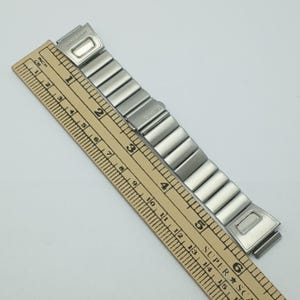 Puede incluir: Una correa de reloj Casio plateada con eslabones rectangulares. La correa se mide con una regla de madera, mostrando una longitud de aproximadamente 15 cm. El logotipo de Casio es visible.