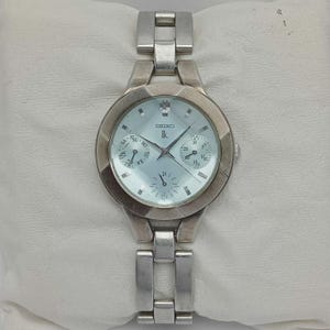 May include: A silver Seiko Ik wristwatch with a light blue face. The watch features a date display, day of the week display, and a silver link band. The watch is displayed on a white surface.