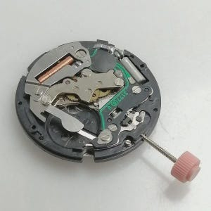 Puede incluir: Primer plano de un movimiento de reloj con engranajes intrincados, circuitos y componentes met&aacute;licos. El mecanismo del reloj es negro con detalles plateados y cobrizos. Una corona rosa est&aacute; adjunta. La imagen destaca la precisi&oacute;n de la relojer&iacute;a.