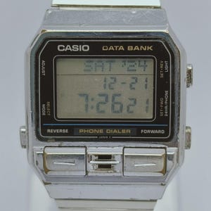 Pode incluir: Um relógio digital Casio Data Bank vintage com caixa e pulseira de metal prateado. O mostrador do relógio exibe a data e a hora. O relógio tem vários botões e o texto "PHONE DIALER".