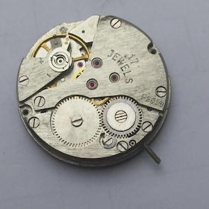 May include: Close-up of a vintage watch movement with visible gears, jewels, and screws. The metal components are silver-toned, with gold and red accents. The text "JEWELS 17" and "P2659" are printed on the surface. A small screw extends from the side.
