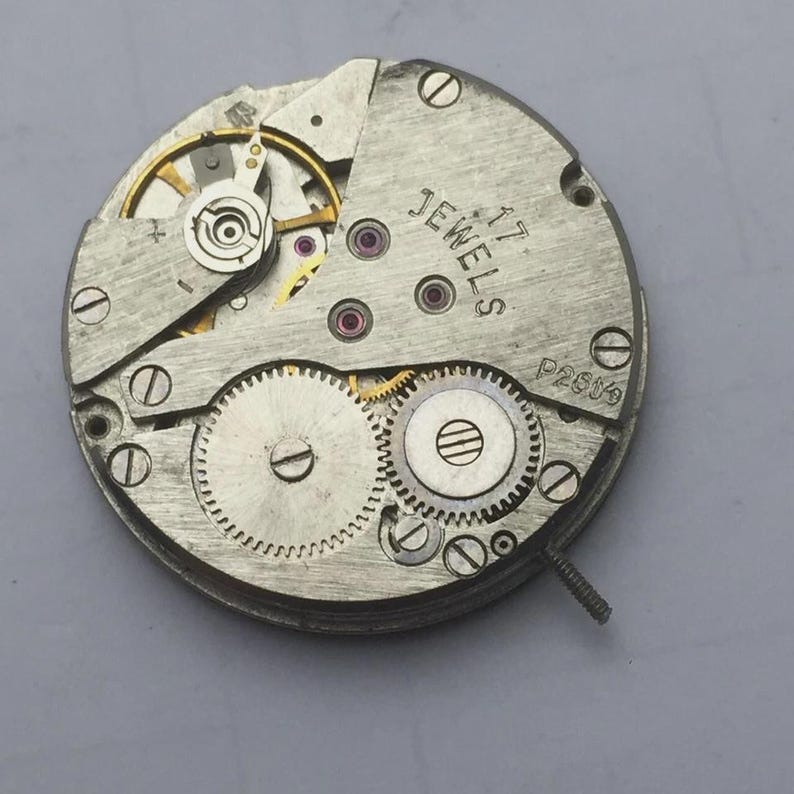 May include: Close-up of a vintage watch movement with visible gears, jewels, and screws. The metal components are silver-toned, with gold and red accents. The text "JEWELS 17" and "P2659" are printed on the surface. A small screw extends from the side.