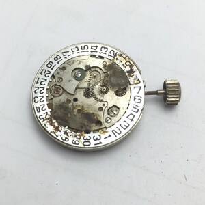 Pu&ograve; includere: Primo piano di un movimento di orologio con ingranaggi visibili, un quadrante della data e una corona. I componenti metallici mostrano segni di usura. Il quadrante della data &egrave; contrassegnato con numeri da 1 a 31. La corona &egrave; a destra.