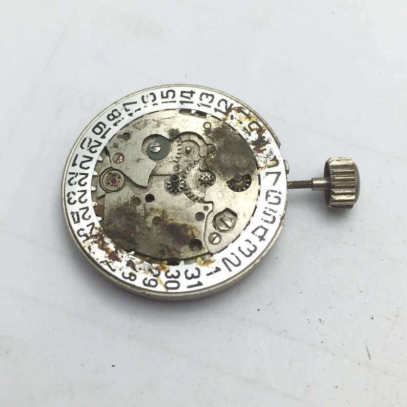 Pu&ograve; includere: Primo piano di un movimento di orologio con ingranaggi visibili, un quadrante della data e una corona. I componenti metallici mostrano segni di usura. Il quadrante della data &egrave; contrassegnato con numeri da 1 a 31. La corona &egrave; a destra.