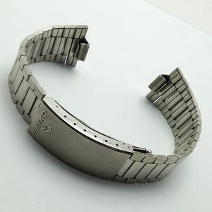 以下が含まれることがあります： 銀色のセイコー腕時計バンド。アーチ状に湾曲しており、折りたたみ式の留め金が付いています。バンドは小さな長方形の金属リンクでできています。セイコーのロゴが留め金に表示されています。このバンドは腕時計に合うように設計されています。