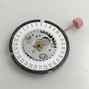 Puede incluir: Primer plano de un movimiento de reloj con una esfera blanca con n&uacute;meros negros del 1 al 31. El movimiento tiene un mecanismo de engranajes plateado en el centro y una corona rosa. El anillo exterior es negro.