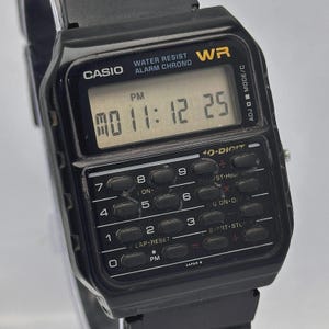 May include: Black Casio digital watch with calculator buttons. The watch face displays the time and date. Features include water resistance and an alarm function. The watch band is black.
