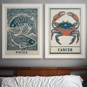 Puede incluir: Dos impresiones artísticas enmarcadas con signos del zodiaco. Una impresión muestra dos peces azules con la palabra "PISCES" debajo. La otra muestra un cangrejo naranja con la palabra "CANCER" debajo. Ambas impresiones tienen un fondo crema.