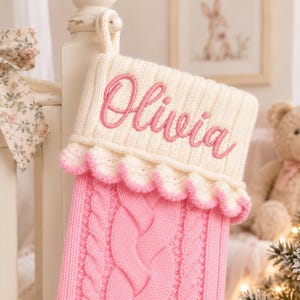 May include: A pink cable-knit Christmas stocking featuring a cream-coloured cuff with the name Olivia embroidered in pink script. The cuff is accented with a delicate pink scalloped trim, and the stocking includes a loop for hanging at the top.