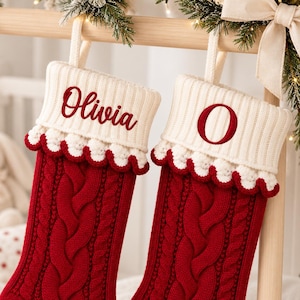 Op de afbeelding: Twee rode kerstsokken van kabelgebreid materiaal met crèmekleurige geribbelde boorden. De linker sok heeft de geborduurde naam Olivia in rood schrift, de rechter sok de rode geborduurde letter O. Beide hebben een geschulpte rand.