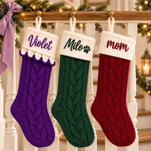 Könnte beinhalten: Drei Weihnachtsstrümpfe aus Zopfstrick hängen an einem Holzgeländer. Der linke Strumpf ist lila mit einer weißen Manschette, die mit dem Namen Violet bestickt ist. Der mittlere ist grün mit einer weißen Manschette, dem Namen Milo und einem Pfotenabdruck. Der rechte ist rot mit einer weißen Manschette, die mit dem Wort mom bestickt ist.