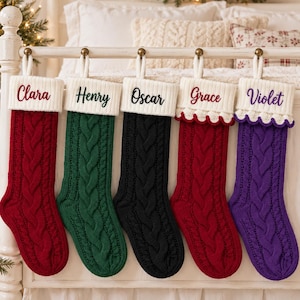 Op de afbeelding: Vijf kerstsokken van kabelbreisel hangen op een rij. De sokken zijn rood, groen, zwart, rood en paars. Elke sok heeft een witte boord geborduurd met de namen Clara, Henry, Oscar, Grace en Violet in verschillende kleuren garen.