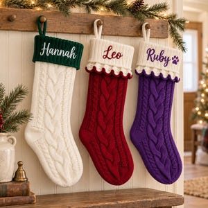 Könnte beinhalten: Drei personalisierte Weihnachtsstrümpfe mit Zopfstrickmuster. Der erste ist weiß mit einem dunkelgrünen Umschlag, bestickt mit "Hannah". Der zweite ist rot mit einem weißen Umschlag, bestickt mit "Leo". Der dritte ist lila mit einem weißen Umschlag, bestickt mit "Ruby" und einem Pfotenabdruck.
