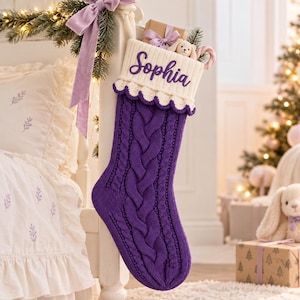 Op de afbeelding: Een dieppaarse kerstkous van kabelgebreid materiaal met een witte geribbelde boord met een geschulpte rand. De naam Sophia is in paars cursief schrift op de boord geborduurd. De kous is gevuld met een kleine knuffel en een zuurstok.
