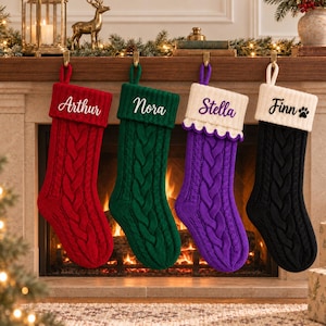 Op de afbeelding: Vier kerstsokken van kabelbreisel hangen op een rij. De eerste is rood met de naam Arthur, de tweede is groen met Nora, de derde is paars met een witte schulprand en de naam Stella, en de vierde is zwart met de naam Finn.