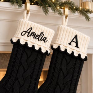 Könnte beinhalten: Zwei schwarze Weihnachtsstrümpfe aus Zopfstrick mit einem cremefarbenen Rippbündchen und gewelltem Rand. Ein Strumpf ist mit dem Namen Amelia in schwarzer Schreibschrift bestickt, der andere zeigt den Buchstaben A in einer schwarzen Serifenschrift.
